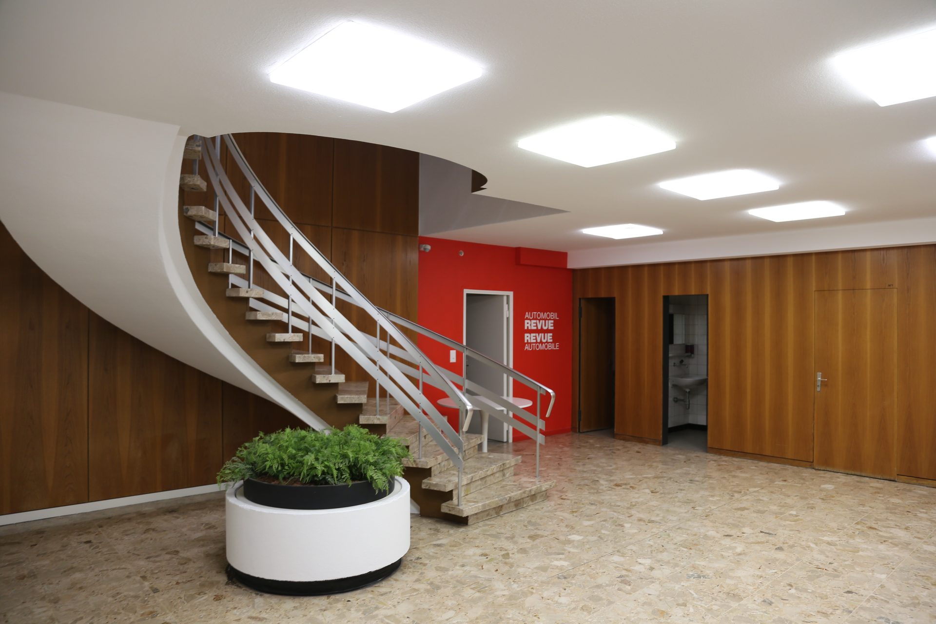 Foyer mit geschwungener Treppe, Stahlgeländer und roter Wand mit Schriftzug «Automobil Revue»