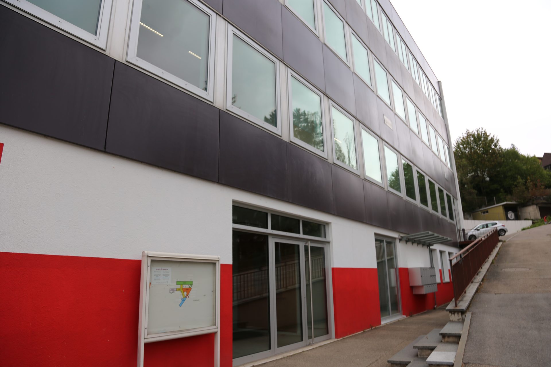 Aussenansicht des Praxisgebäudes mit grau-weiss-roter Fassade, Glaseingang und seitlicher Rampe