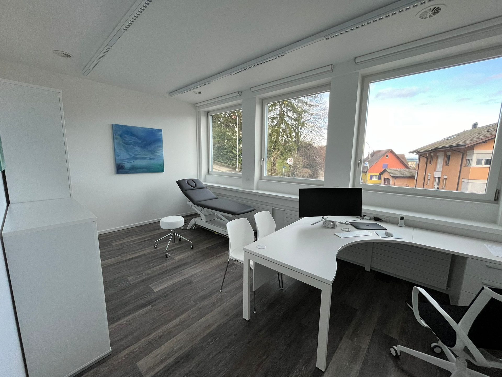 Helles Sprechzimmer der Praxis mit Behandlungsliege, Schreibtisch und grossen Fenstern